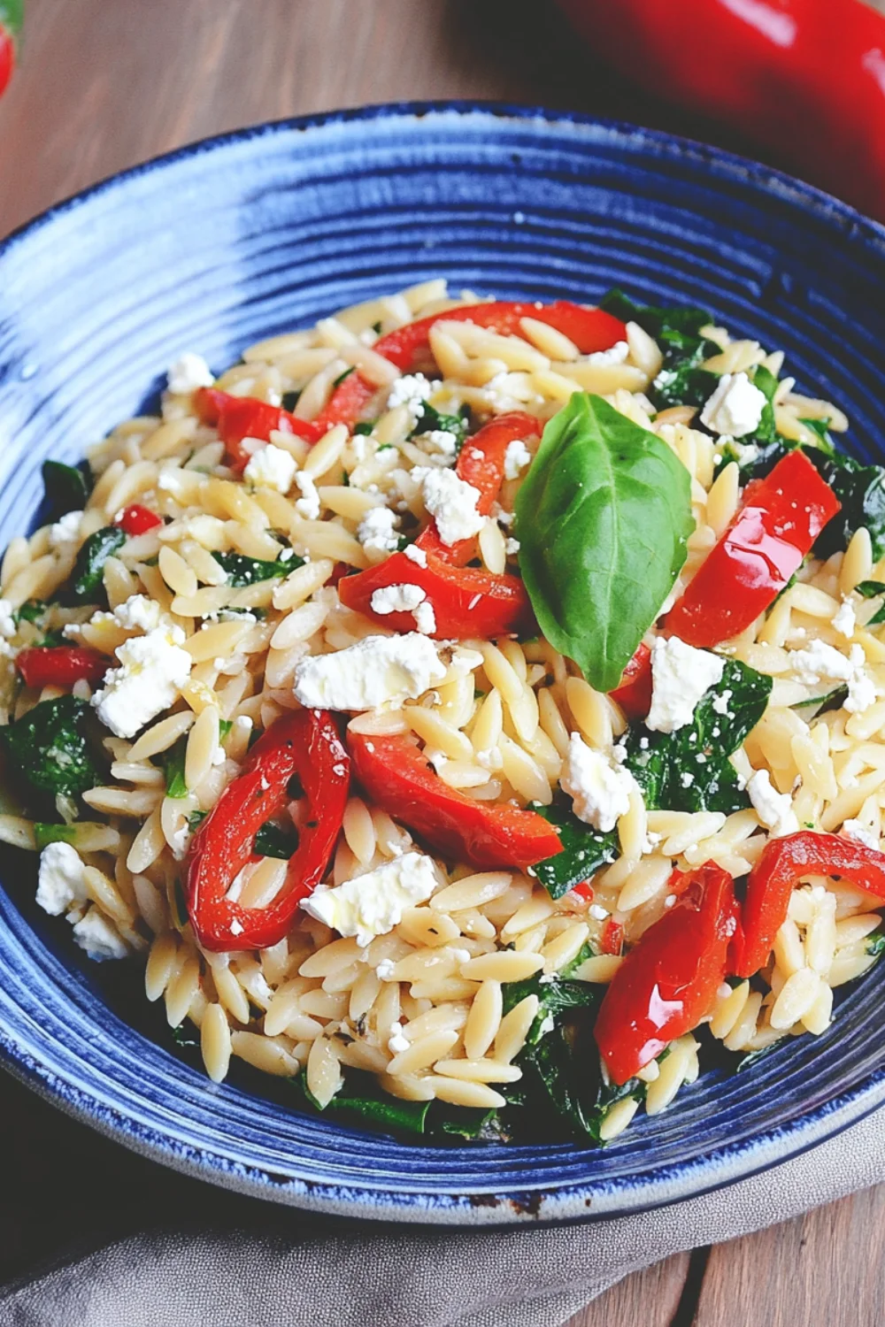Orzo-Nudelsalat mit gerösteten roten Paprikastreifen, frischem Spinat und zerbröseltem Feta in einer blauen Schale, garniert mit einem Basilikumblatt.